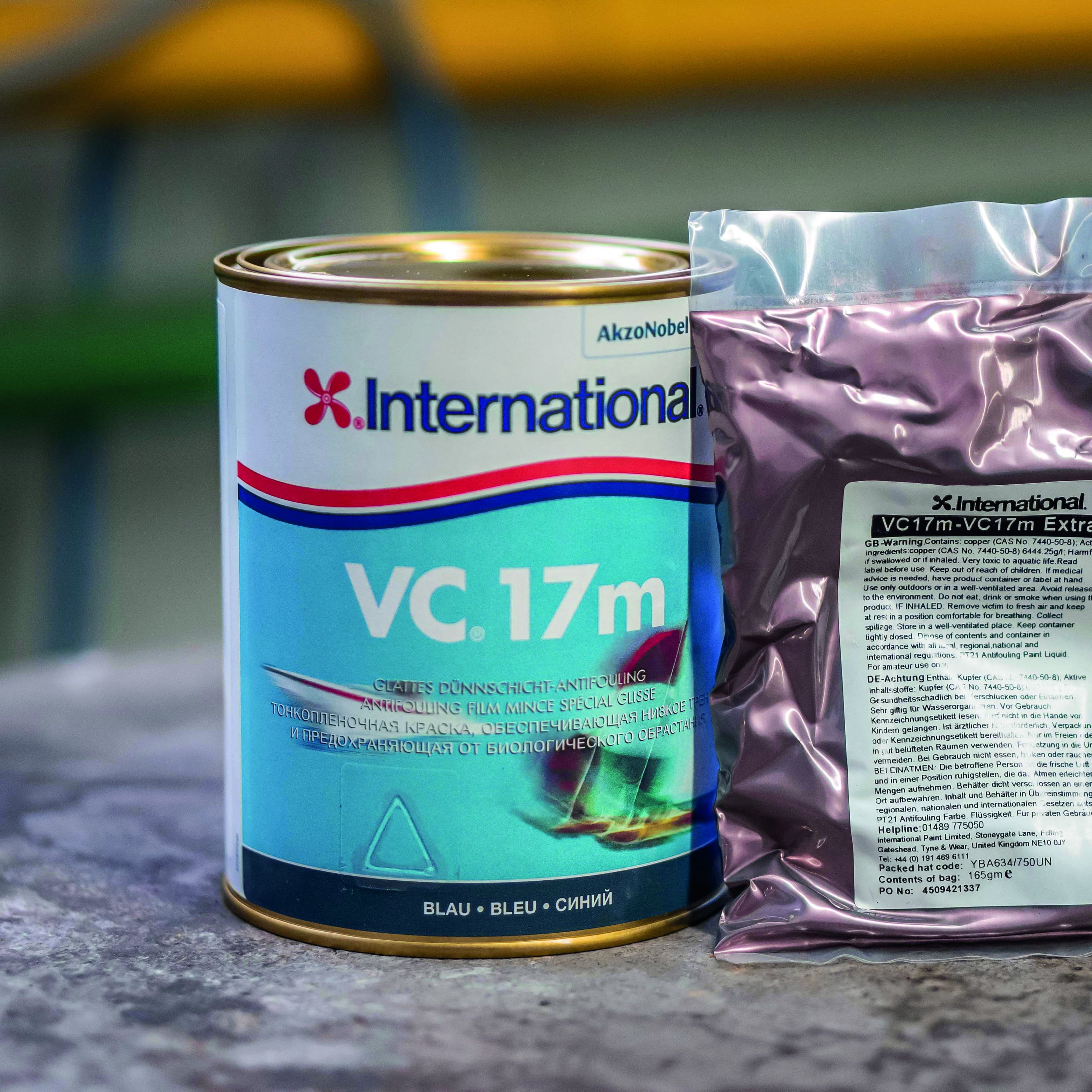 Eine Dose mit der Aufschrift "International VC 17m" in Blau und eine Verpackung mit violettem Inhalt. Die Dose zeigt ein Schiffssymbol und Informationen zur Verwendung des Produkts, während die Verpackung eine klare Anleitung auf der Rückseite hat. Im Hintergrund sind unscharfe, grüne und gelbe Elemente zu erkennen.