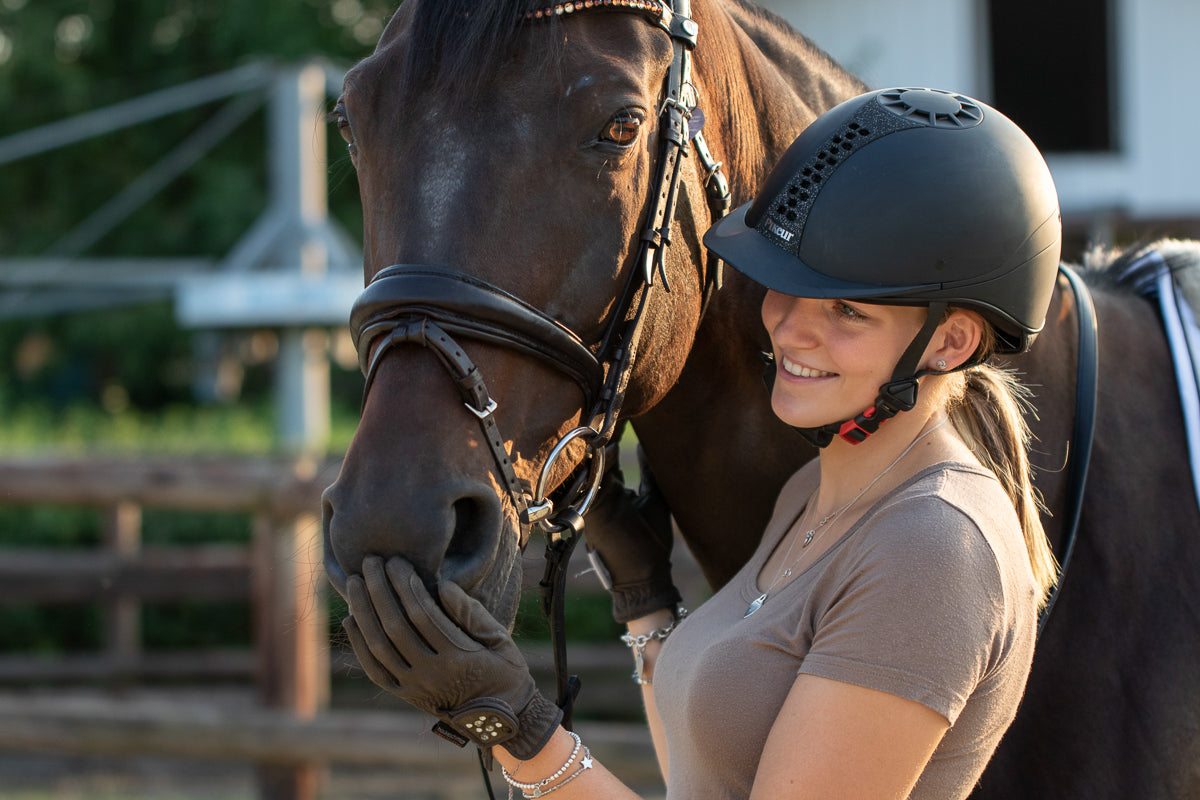 Eine junge Frau mit einem Schutzhelm und Reithandschuhen lächelt, während sie liebevoll mit einem großen, braunen Pferd interagiert. Das Pferd trägt ein Kopfstück und steht ruhig. Im Hintergrund sind Holzbarrieren und eine Weide zu sehen, die eine ländliche Umgebung schaffen. Die Abendsonne beleuchtet die Szene und sorgt für eine warme, freundliche Atmosphäre.
