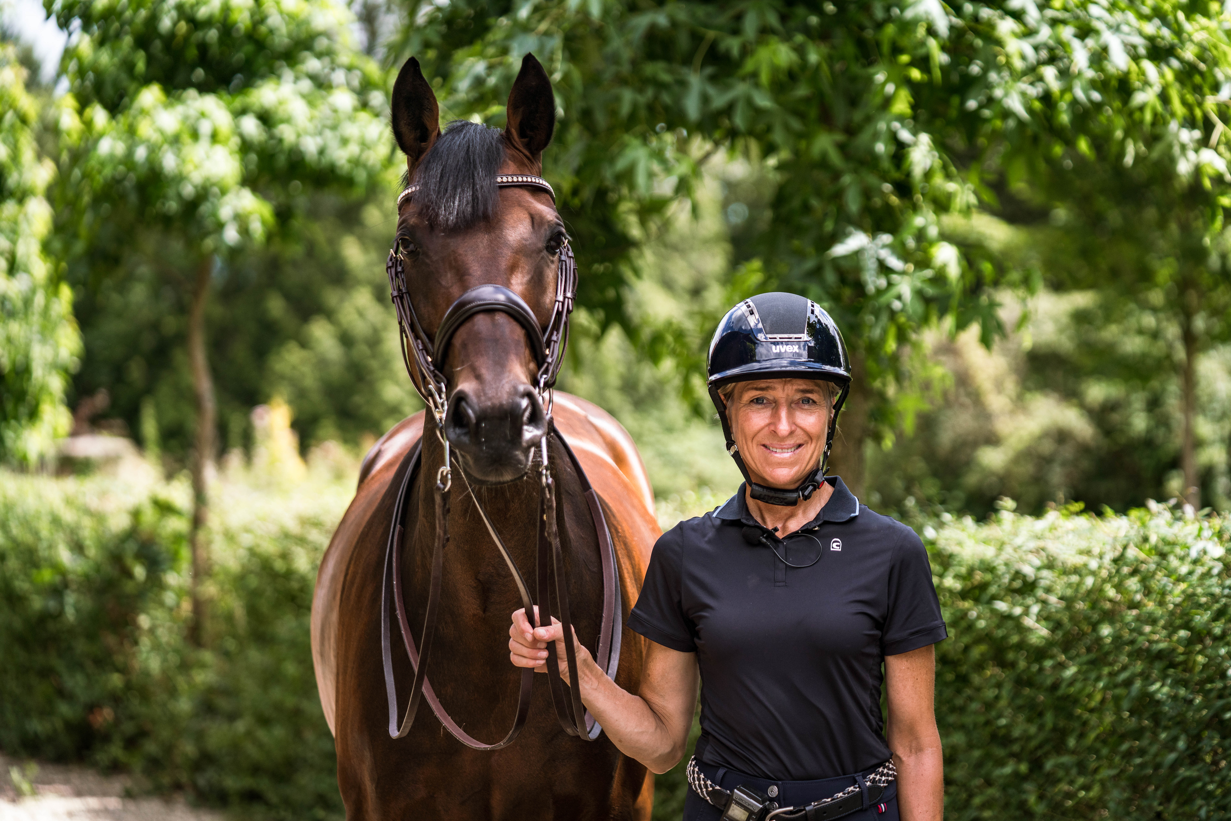 Eine reitende Person in einem schwarzen Shirt und Helm steht neben einem braunen Pferd. Die Person lächelt und hält das Zügel des Pferdes. Im Hintergrund sind grüne Bäume und eine gepflegte Hecke zu sehen, die eine natürliche Umgebung schaffen. Das Bild vermittelt eine freundliche, harmonische Stimmung zwischen Mensch und Tier.