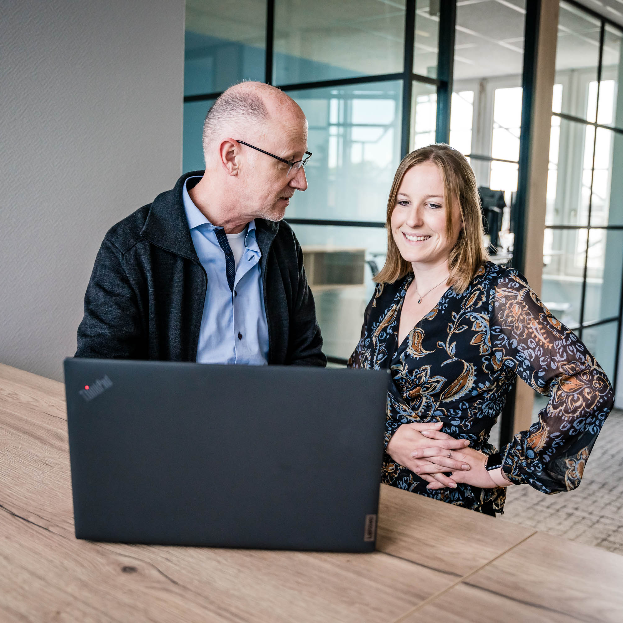 Zwei Personen sitzen an einem Tisch in einem modernen Büro. Ein Mann und eine Frau schauen gemeinsam auf einen Laptop. Der Mann trägt eine schwarze Jacke und eine blaue Bluse, während die Frau ein blütenreiches, farbiges Oberteil trägt. Beide wirken engagiert und unterhalten sich freundlich. Im Hintergrund sind große Fenster, die Tageslicht hineinlassen.
