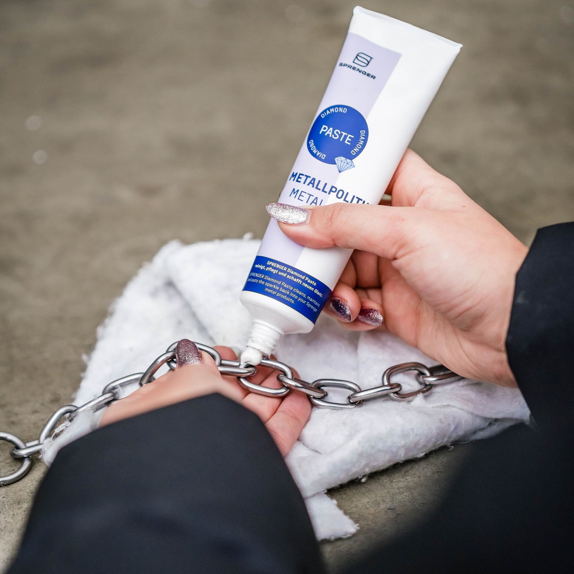 Eine Hand hält eine Tube Metallpolitur mit einem weißen Etikett, während eine zweite Hand ein Reinigungstuch mit einer Kette daneben hält. Der Hintergrund zeigt einen grauen Bodenbelag.