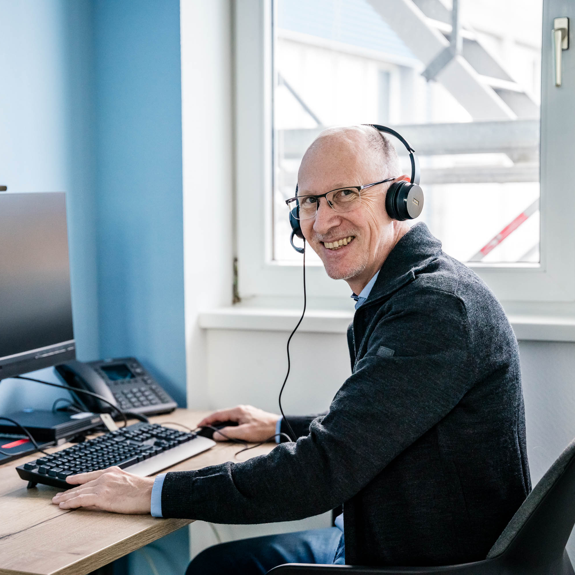 Ein älterer Mann mit Brille sitzt an einem Schreibtisch vor einem Computerbildschirm. Er trägt ein Headset und lächelt in die Kamera. Auf dem Tisch sind zudem eine Tastatur und ein Telefon sichtbar. Der Raum hat ein helles und modernes Ambiente mit einem Fenster, durch das man eine Fassade und Gerüst sehen kann.