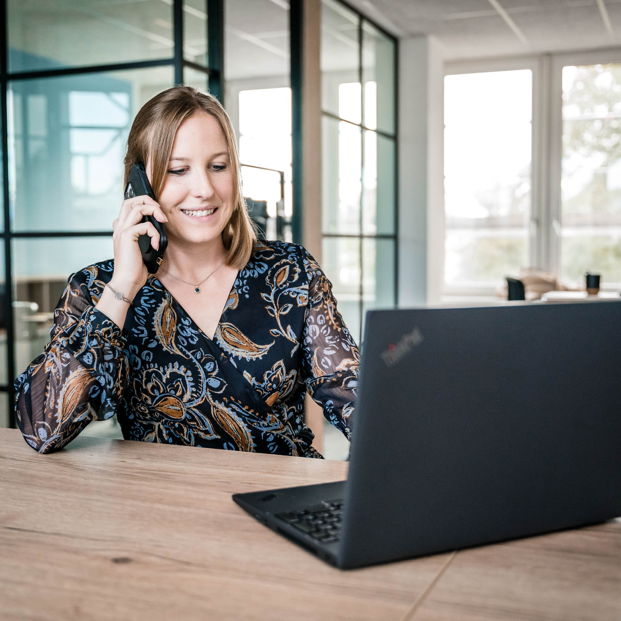 Eine lächelnde Frau mit langen, blonden Haaren sitzt an einem holzfarbenen Tisch in einem modernen Büro. Sie spricht telefonierend und sieht auf einen Laptop, der geöffnet vor ihr steht. Im Hintergrund sind große Fenster zu sehen, die Tageslicht hereinlassen. Die Frau trägt ein dunkelblaues, gemustertes Oberteil.