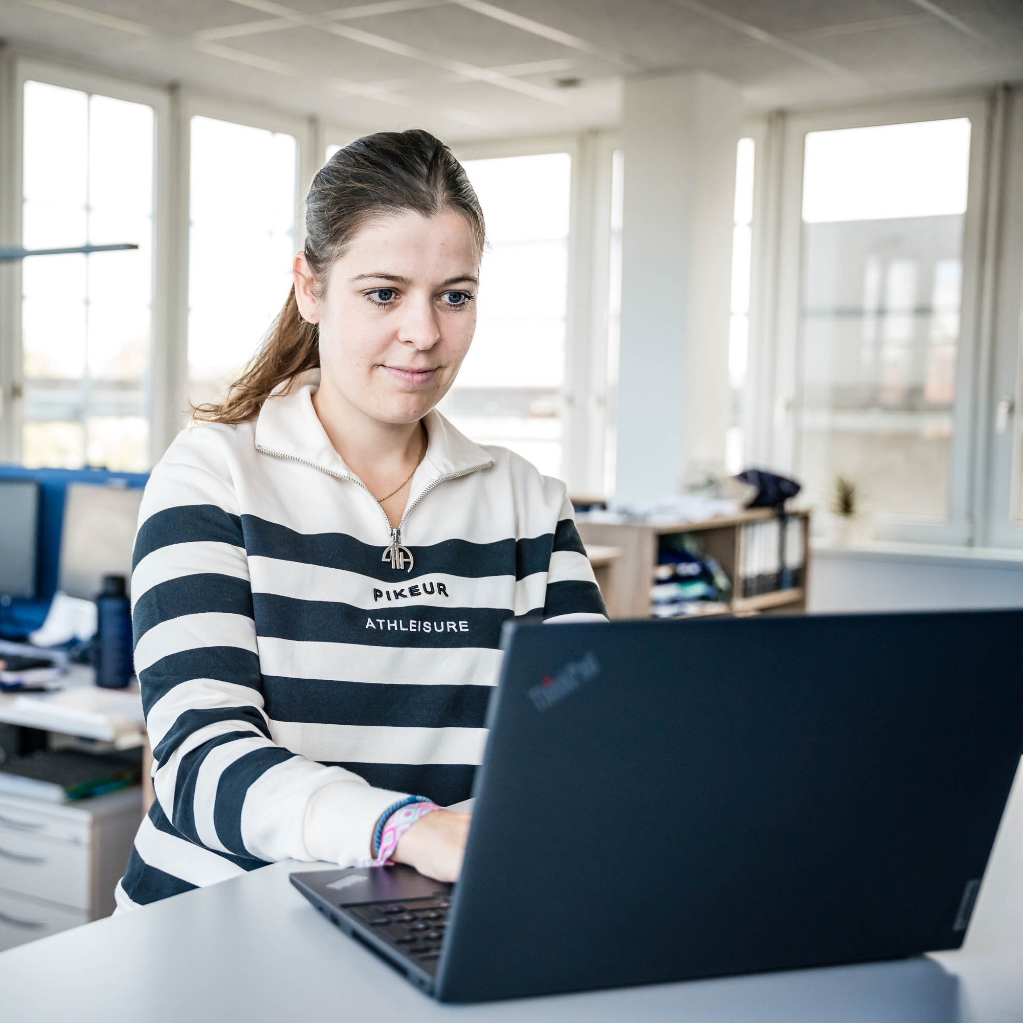Eine junge Frau mit langen, braunen Haaren, die zu einem Pferdeschwanz gebunden sind, sitzt an einem Schreibtisch und arbeitet an einem Laptop. Sie trägt einen gestreiften Pullover in Schwarz und Weiß mit dem Schriftzug "Pikeur Athleisure". Im Hintergrund sind Büromöbel und Fenster mit natürlichem Licht zu sehen, was eine helle und freundliche Büroatmosphäre vermittelt.
