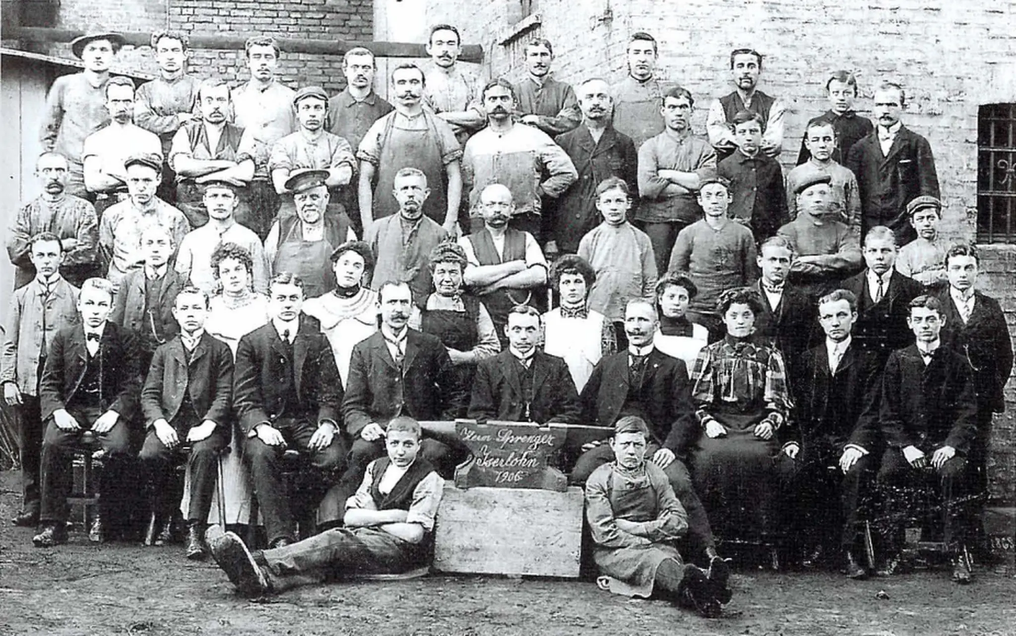 Gruppenfoto aus der Vergangenheit, das eine Vielzahl von Menschen zeigt, darunter Männer und Frauen unterschiedlicher Alter. Die Personen stehen und sitzen in mehreren Reihen vor einer Backsteinwand. Einige tragen historische Arbeitskleidung, während andere elegant gekleidet sind. In der Mitte des Bildes steht ein Stein mit der Aufschrift "Jahresfeier 1916". Die Atmosphäre vermittelt ein Gefühl von Gemeinschaft und Feierlichkeit. Das Bild ist in Schwarz-Weiß und vermittelt einen nostalgischen Eindruck.