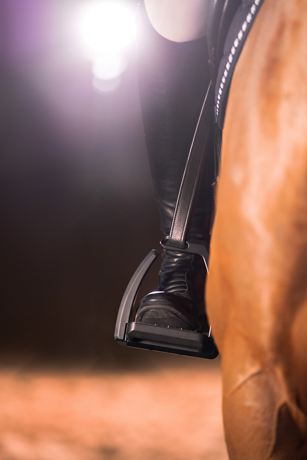 Nahaufnahme eines Reiters, der in einem Steigbügel sitzt. Der Fokus liegt auf dem schwarzen Steigbügel und dem rechten Fuß des Reiters, der bottschen zur Seite zeigt. Der Hintergrund ist verschwommen mit weichem Licht, das von einer Quelle oben links strahlt, was eine stimmungsvolle Atmosphäre schafft. Der Pferdekörper ist ebenfalls teilweise sichtbar, was die Reitsituation unterstreicht.