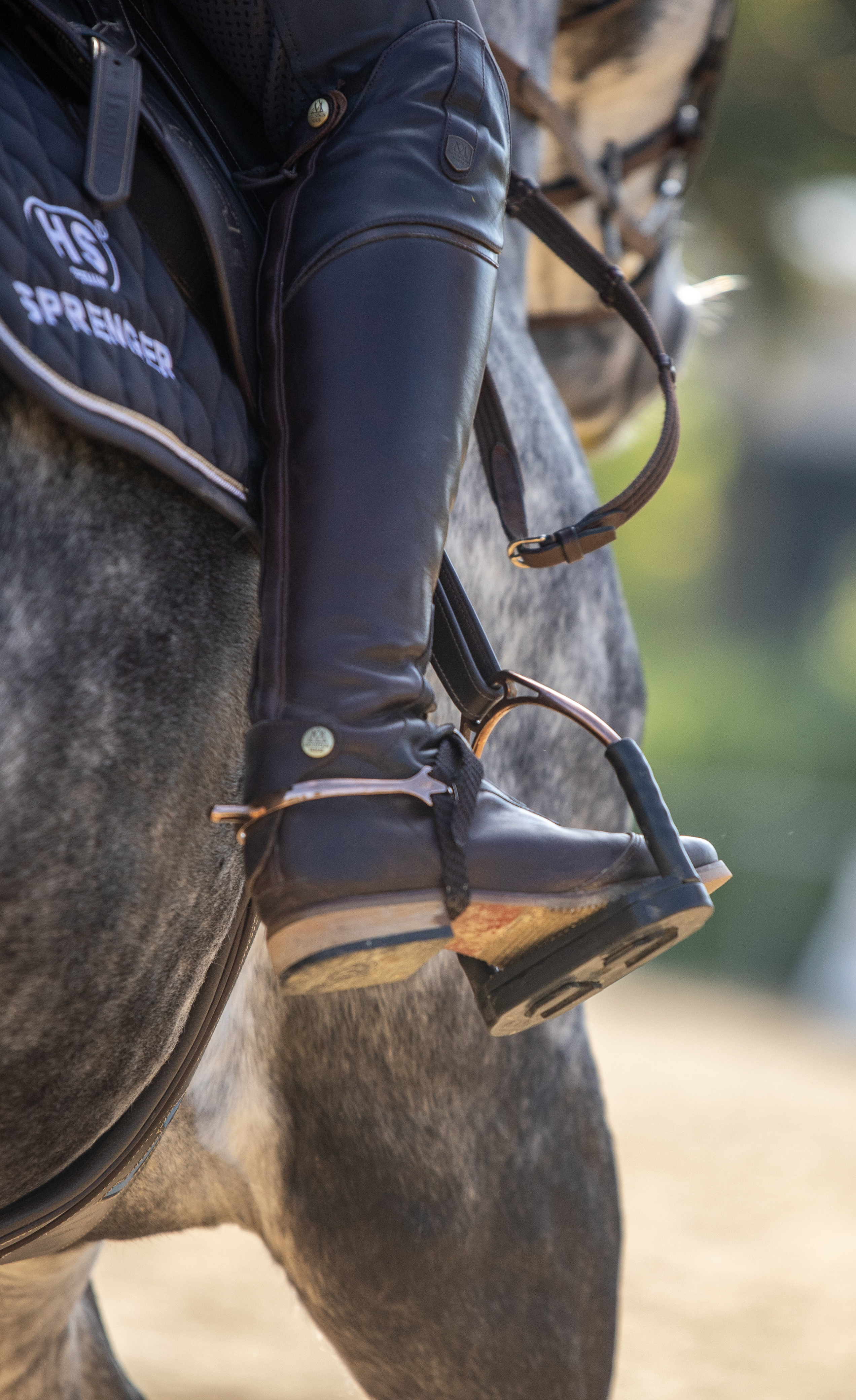 Nahaufnahme eines Reiters mit schwarzem Stiefel, der im Steigbügel sitzt. Der Stiefel ist aus Leder und zeigt eine seitliche Ansicht. Im Hintergrund ist ein graues Pferd zu erkennen, sowie der Sattel und der Platz, auf dem geritten wird.