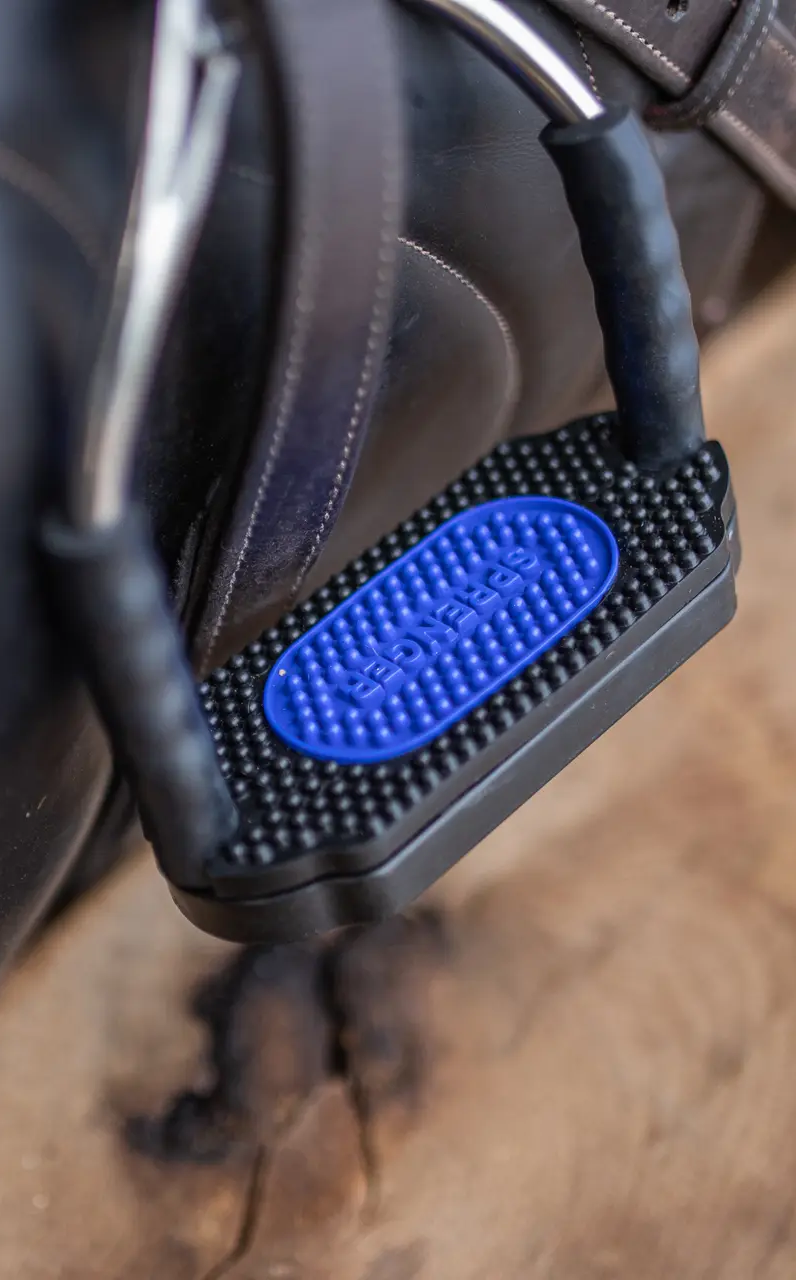 Ein nahe aufgenommenes Bild eines Sattelsteigs aus schwarzem Kunststoff mit blauer Gummieinlage. Die rutschfeste Oberfläche hat eine strukturierte Form, die für besseren Halt sorgt. Der Steigbügel ist an einem braunen Sattel befestigt und zeigt die Schnalle, die zur Befestigung dient. Der Hintergrund besteht aus Holz, das die Natürlichkeit des Reiterumfelds betont.