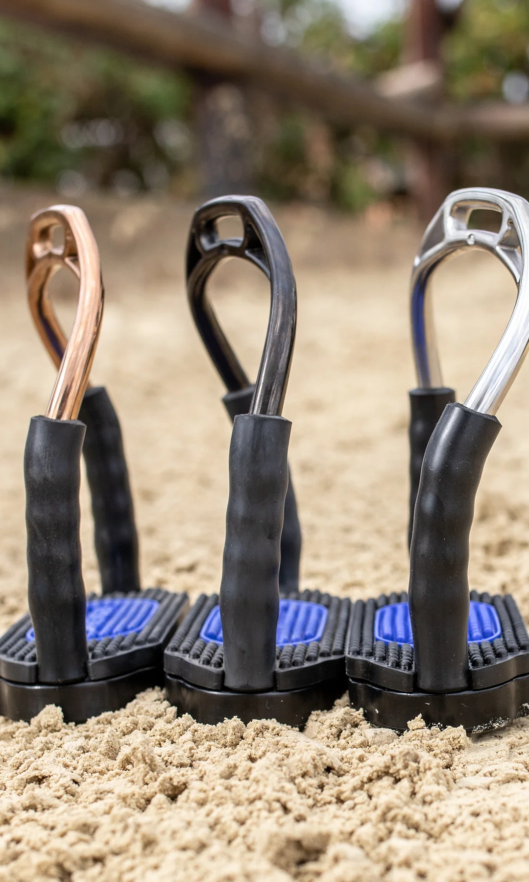 Drei verschiedenfarbige Stäbe mit ergonomischen Griffen stehen auf einem sandigen Untergrund. Die Stäbe haben farbige Kopfbänder in Kupfer, Schwarz und Silber und sind mit blauem Gummi an der Unterseite ausgestattet. Der Fokus liegt auf den Griffen und der Verbindung zur sandigen Fläche.