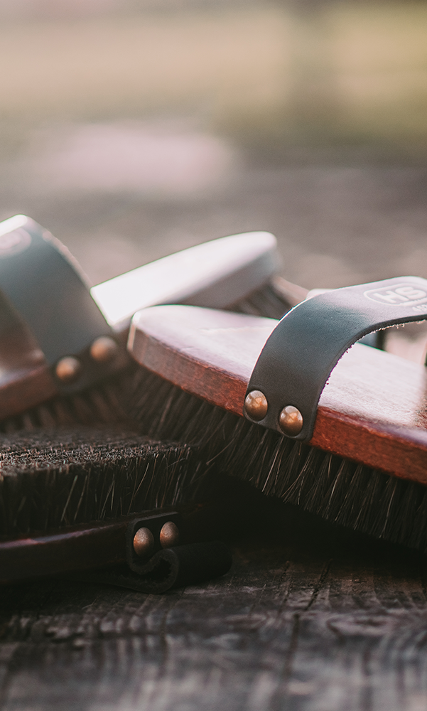 Zwei Holzschuhe mit Bürstensohlen liegen auf einer Holzoberfläche. Die Schuhe haben schwarze Riemen und Metallnieten. Der Hintergrund ist unscharf und in sanften Farben gehalten.