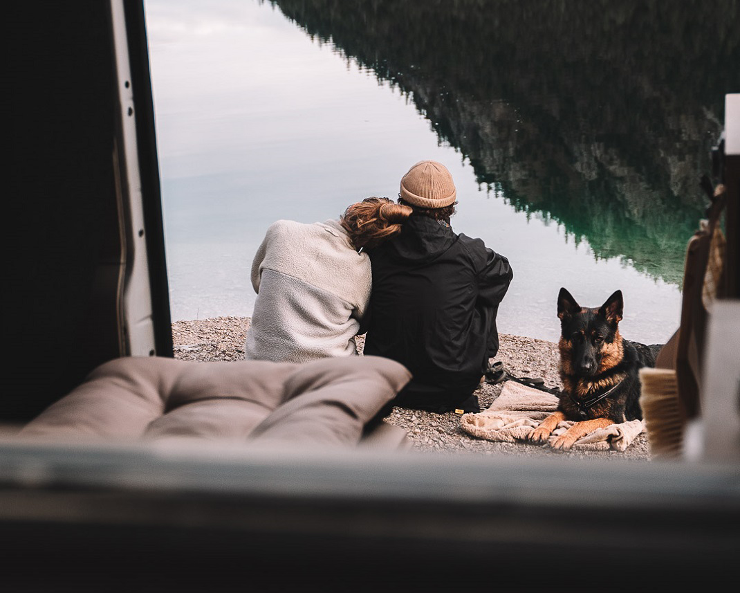 Ein Paar sitzt am Ufer eines ruhigen Sees und blickt auf das Wasser. Die Person in einem weißen Pullover lehnt sich an die andere Person, die einen Hut trägt. Neben ihnen sitzt ein schwarzer Hund. Im Vordergrund ist eine gepolsterte Unterlage zu sehen. Die Umgebungslandschaft ist von Bäumen und Bergen geprägt.