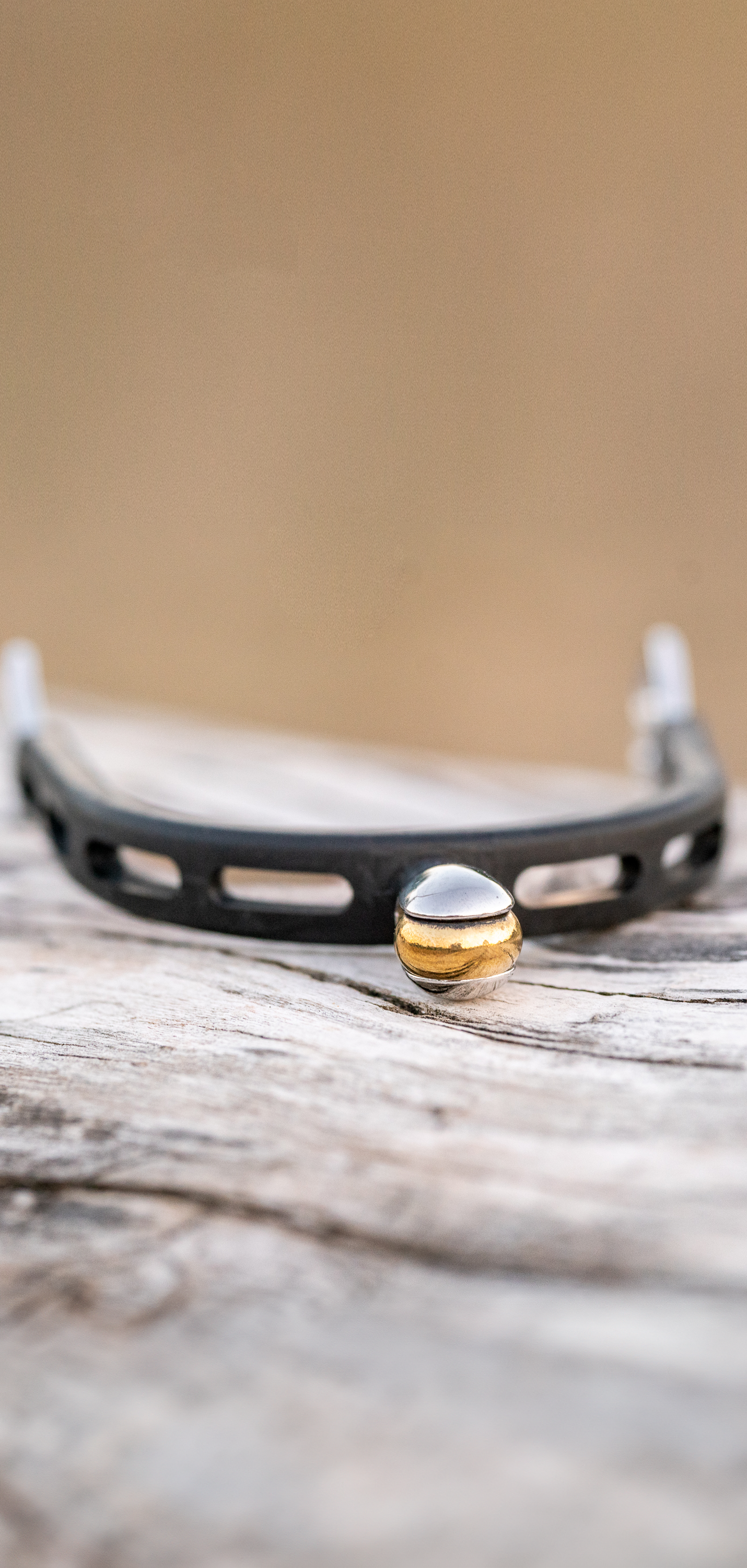 Eine schmale, schwarze Armbanduhr mit abgerundeter, glänzender Uhrkrone aus Gold und Silber liegt auf einem rustikalen Holzuntergrund. Der Fokus liegt auf der Uhr, während der Hintergrund unscharf und in warmen Erdtönen gehalten ist. Das Design der Uhr kombiniert moderne Elemente mit einem Hauch von Eleganz.