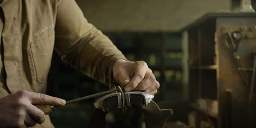 Eine Person in einem braunen Hemd bearbeitet ein Metallteil mit einer Feile. Die Hand ist konzentriert und hält das Werkstück stabil, während die andere Hand die Feile führt. Im Hintergrund sind unscharfe Werkzeuge und Regale zu sehen, die auf eine Werkstatt hinweisen. Die Beleuchtung ist warm und betont die Handwerkskunst.