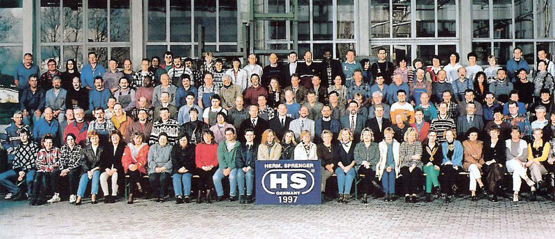 Gruppenfoto von etwa 100 Personen, die in einem großen Raum vor großen Fenstern stehen. Die Menschen sind in verschiedenen Kleidungsstilen gekleidet und lächeln in die Kamera. Im Vordergrund steht ein Schild mit der Aufschrift „Herbal Sprenger HS 1997“. Die Personen sind gemischt zwischen Männern und Frauen und zeigen eine vielfältige kollektive Darstellung.