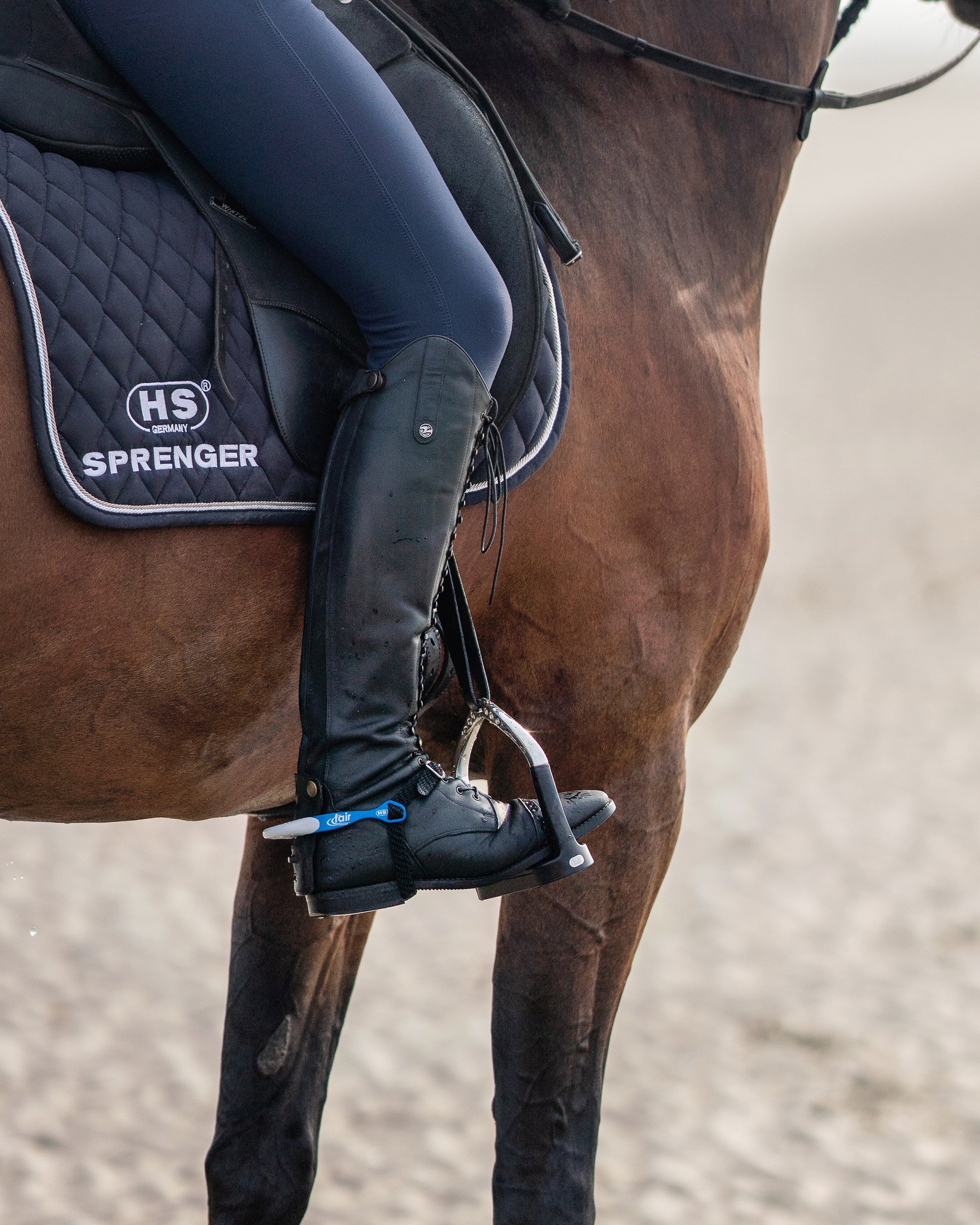 Reiterin, die elegant auf einem braunen Pferd sitzt. Der Fokus liegt auf dem Bein der Reiterin, das in einer schwarzen Reitstiefel steckt und auf dem Steigbügel ruht. Der Sattel ist mit einer schwarzen Sattelunterlage von Sprenger ausgestattet. Im Hintergrund ist ein unscharfer Sandboden zu sehen. Das Bild vermittelt eine ruhige Reitsituation.