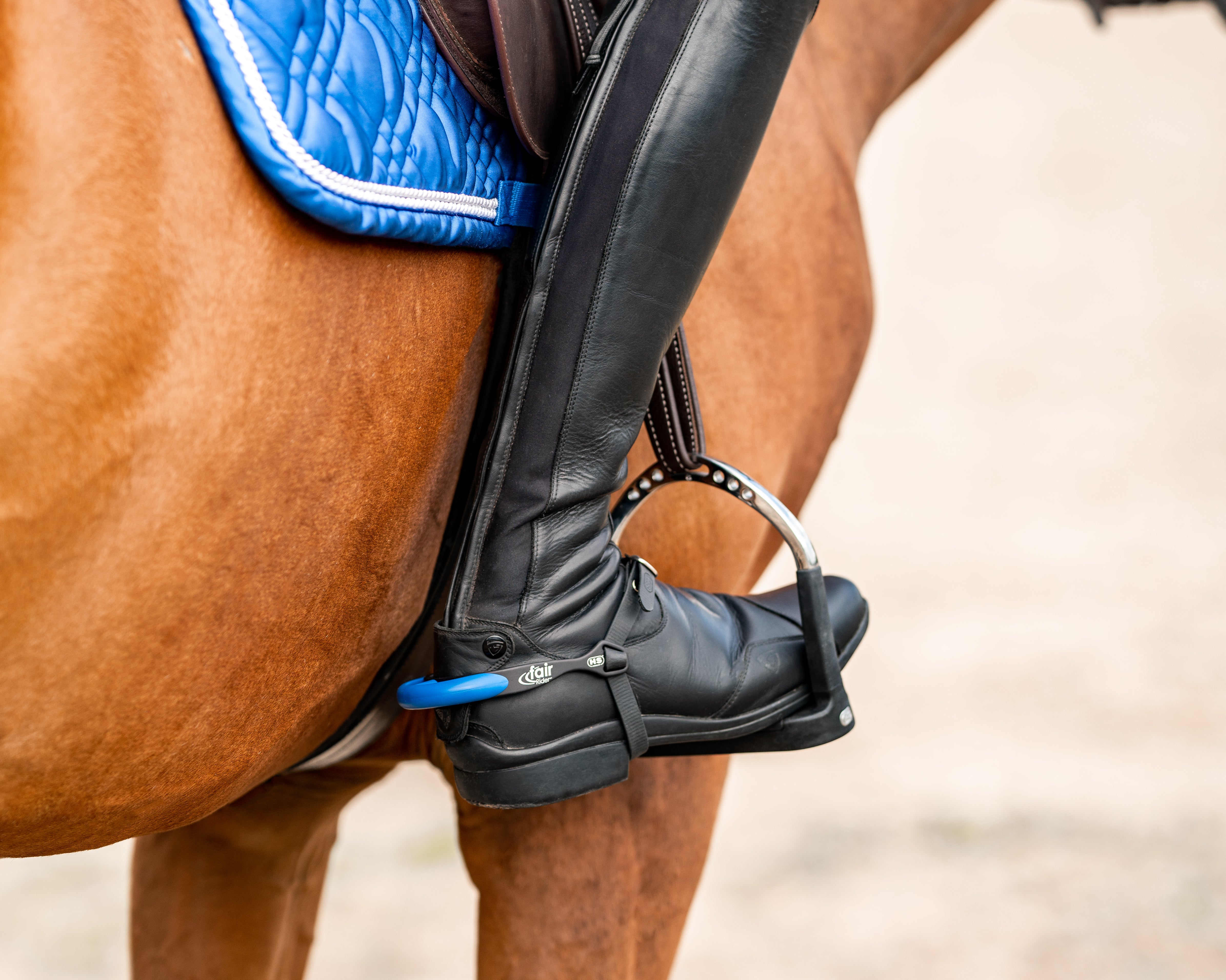Detailaufnahme eines Reiters und eines Pferdes. Im Vordergrund ist ein gefütterter Sattel zu sehen, auf dem ein schwarzer Stiefel sitzt. Der Reiter hat den Fuß im Steigbügel platziert, der mit einem blauen Gurt verbunden ist. Die Umgebung ist sandig und unscharf, während der Fokus auf dem Stiefel und dem Sattel liegt.