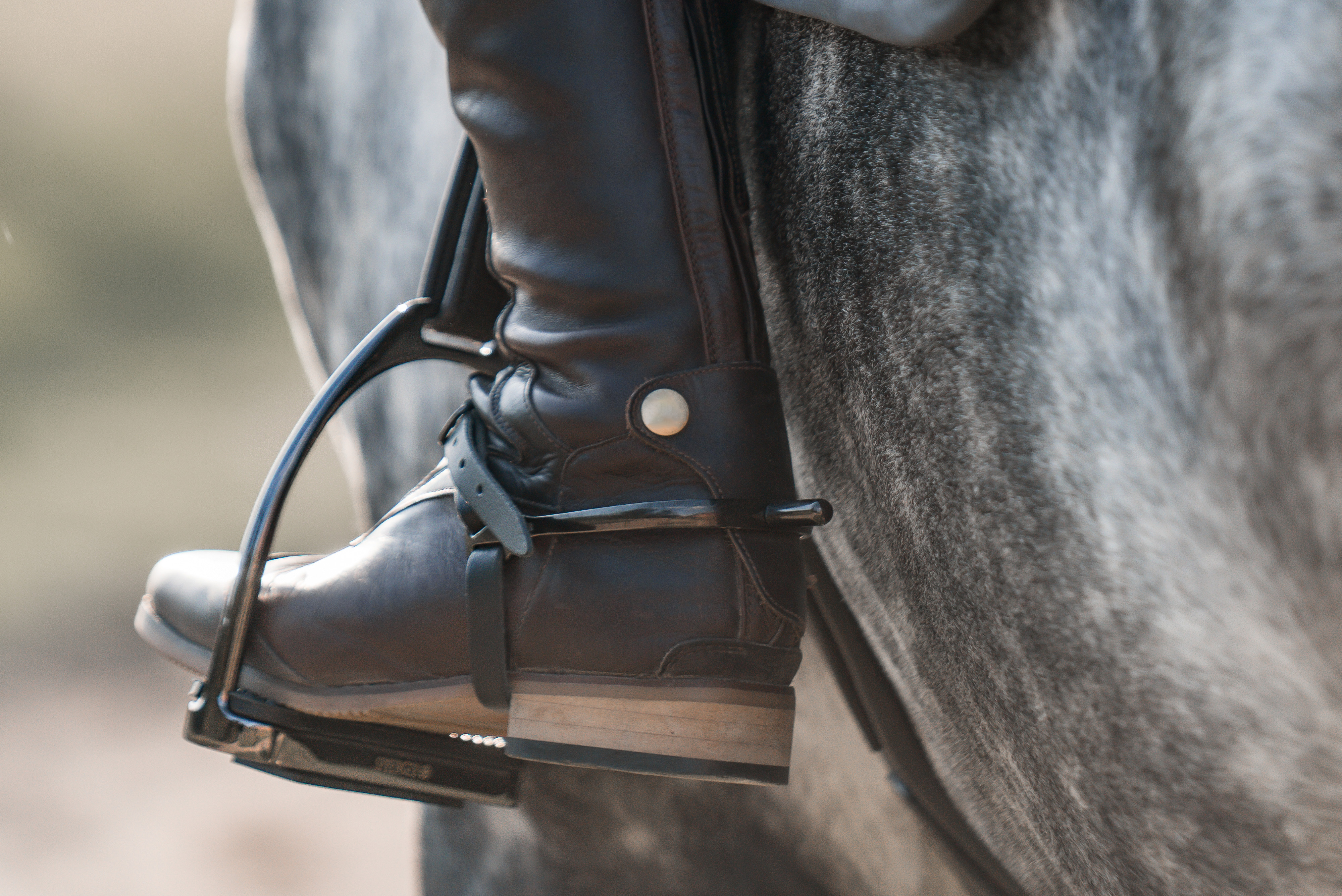 Ein Reiter steht in einem Sattel auf einem grauen Pferd. Der Fokus liegt auf dem rechten Stiefel des Reiters, der in einem Steigbügel steckt. Der Stiefel ist aus braunem Leder und hat einen Holzabsatz. Der Hintergrund ist unscharf, mit einem natürlichen Umfeld.