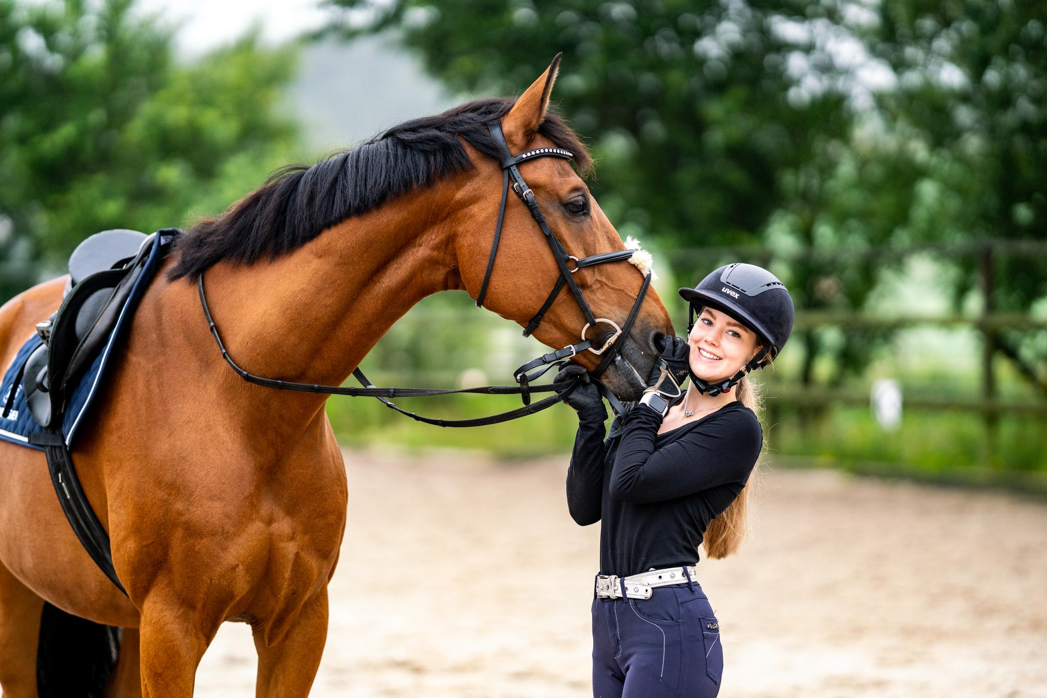 Eine reitende Frau in einem schwarzen Oberteil und einer Reithelmkappe steht vor einem braunen Pferd. Sie lächelt und hält das Pferd sanft am Kopf, während sie ihm in die Augen schaut. Im Hintergrund sind verschwommene, grüne Bäume und ein Reitplatz zu erkennen. Das Bild vermittelt eine freundliche Verbindung zwischen Mensch und Tier.