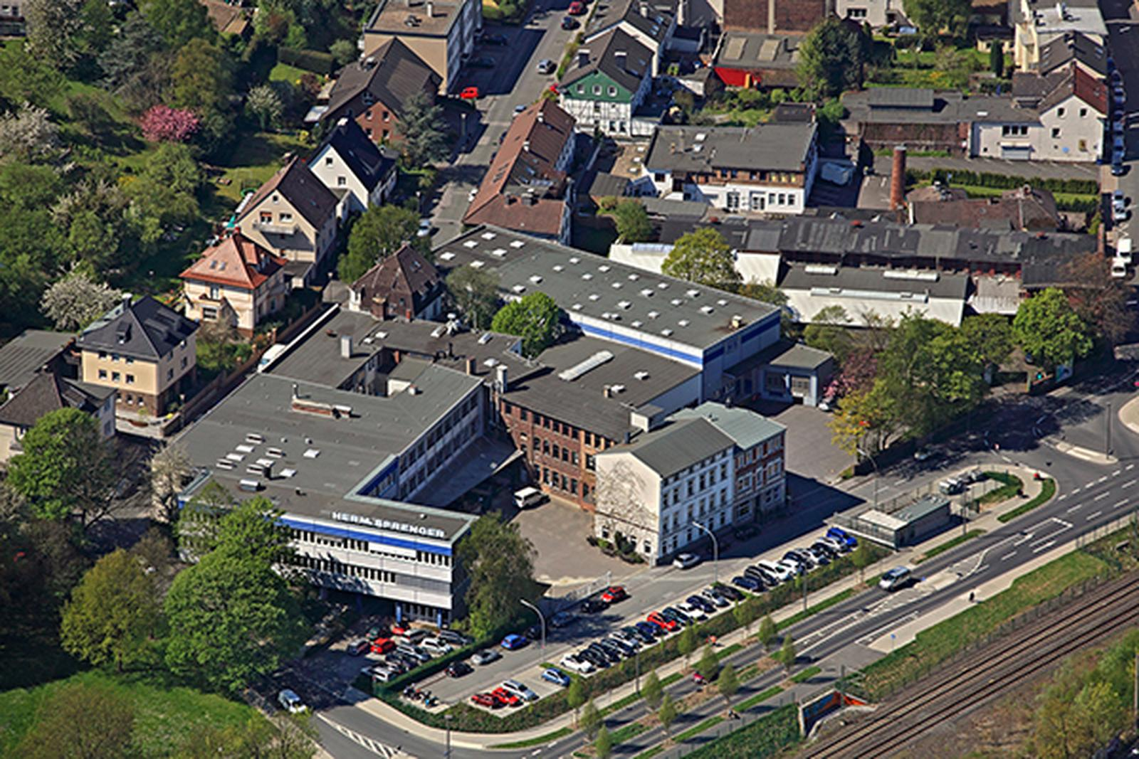 Luftaufnahme eines Stadtgebiets mit mehreren Gebäuden, darunter ein modernes Firmengebäude mit Flachdach und großen Fenstern, sowie ein historisches Backsteingebäude. Die Umgebung ist grün, mit Bäumen und Blumen. Parkplätze sind sichtbar, und eine Straße verläuft entlang des Bildes. Im Hintergrund sind Wohnhäuser und weitere Gebäude der Nachbarschaft zu erkennen.
