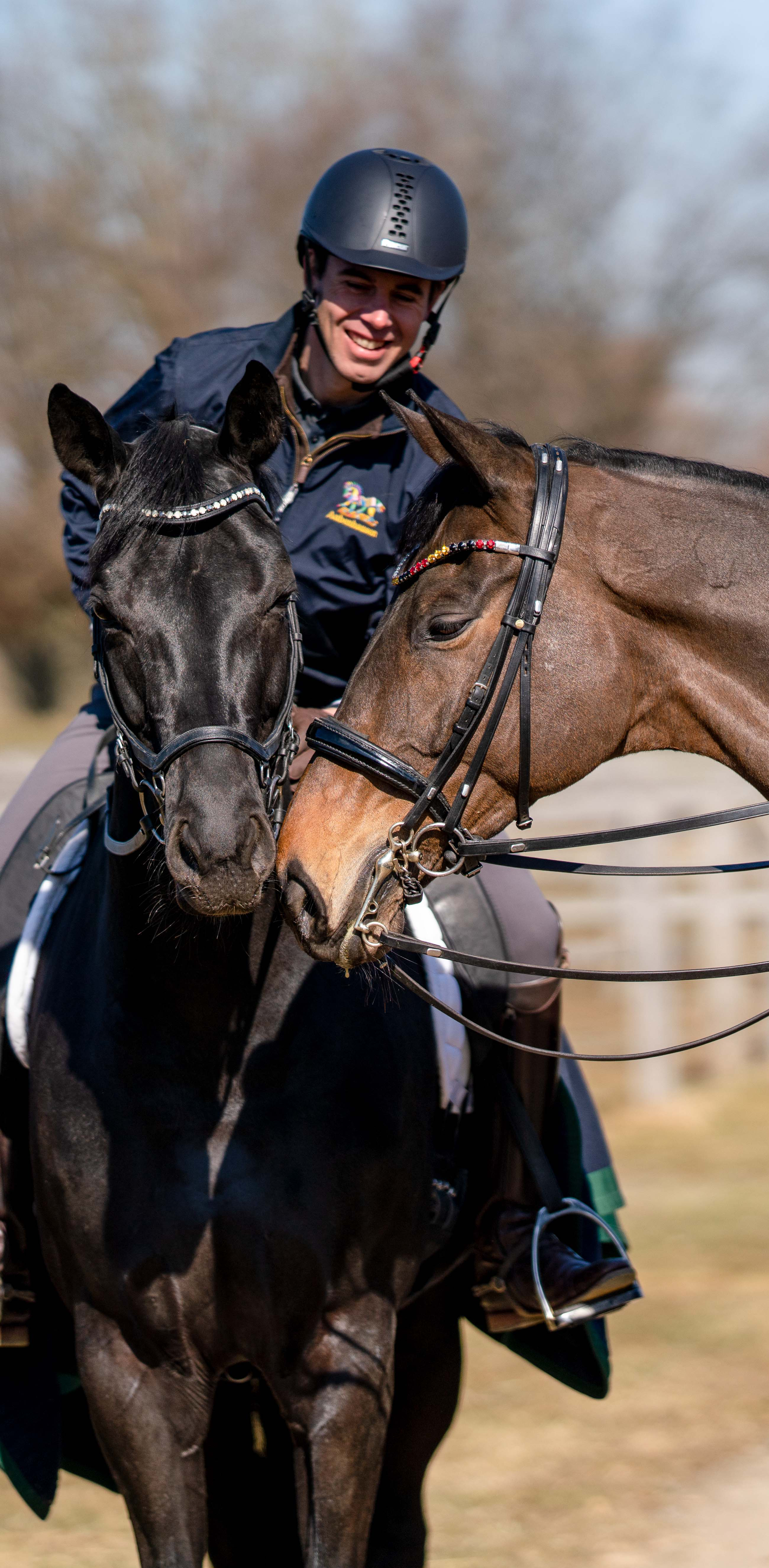 Eine reitende Person mit Helm lächelt glücklich zwischen zwei Pferden, einem schwarzen und einem braunen. Die Pferde stehen dicht beieinander und scheinen sich zu berühren. Die Umgebung ist sonnig und zeigt eine natürliche Landschaft mit Bäumen im Hintergrund. Die Person trägt eine dunkelblaue Jacke mit einem Logo und hat die Zügel der Pferde in der Hand.
