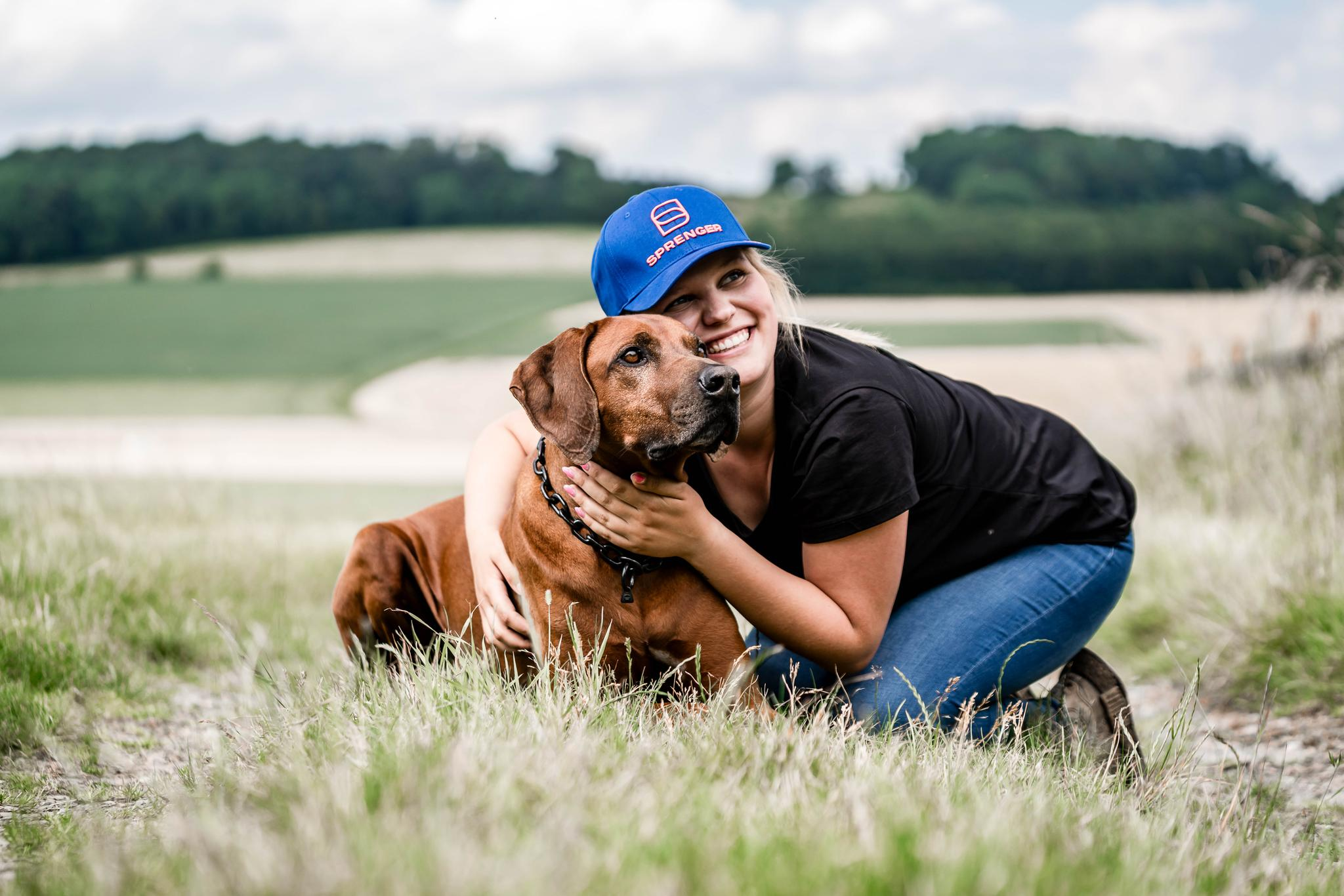 Eine junge Frau mit blonden Haaren, die unter einer blauen Kappe steckt, kniet in einem grasbewachsenen Feld neben einem braunen Hund. Sie umarmt den Hund, während sie lächelt. Im Hintergrund erstrecken sich sanfte Hügel und ein klarer Himmel. Das Bild vermittelt eine freundliche und fröhliche Atmosphäre, die die enge Verbindung zwischen Mensch und Tier zeigt.