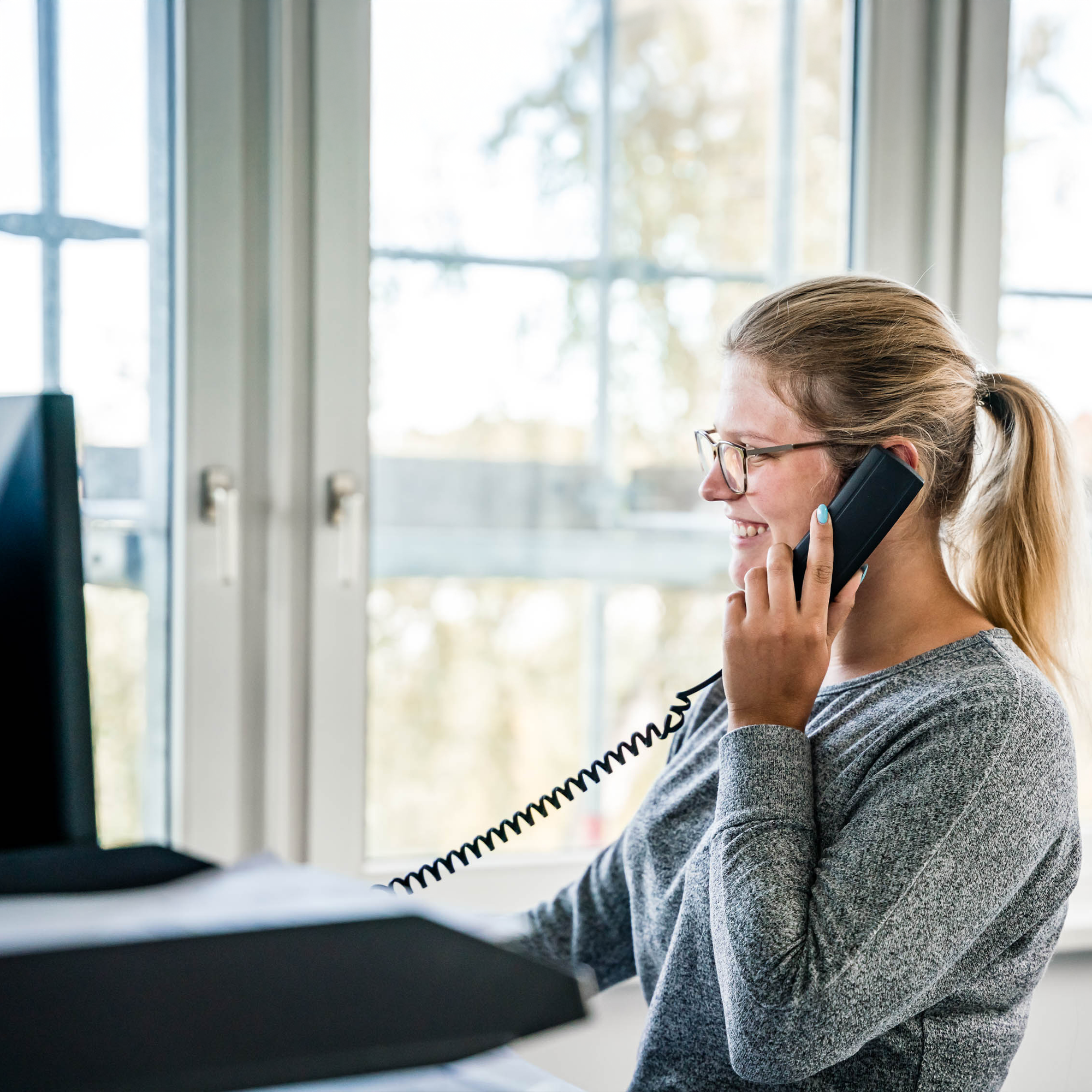 Eine junge Frau mit Brille sitzt an einem Schreibtisch und spricht lächelnd am Festnetztelefon. Im Hintergrund sind große Fenster, durch die Tageslicht strömt. Auf dem Schreibtisch liegt Papier.