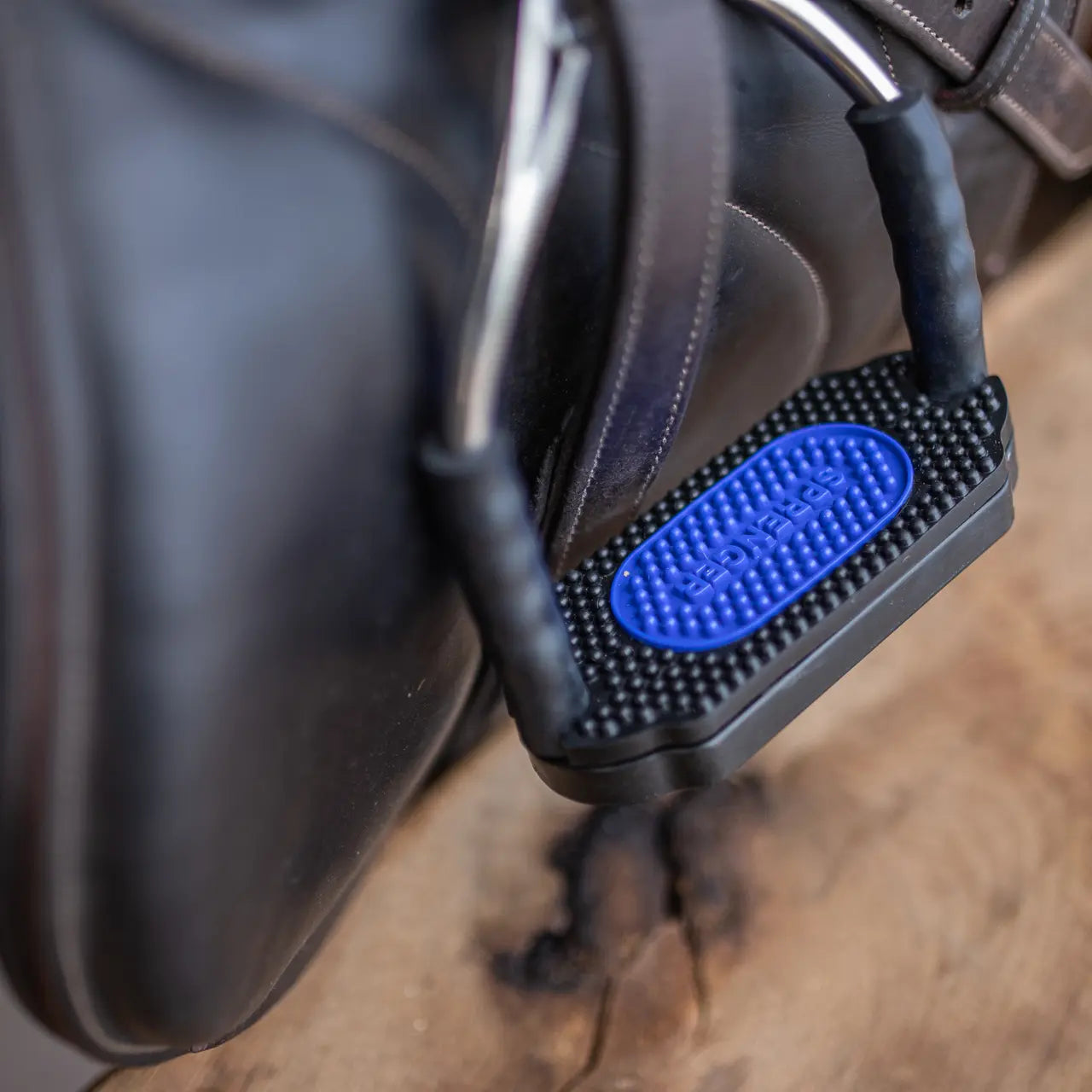 Nahaufnahme eines Sattelgurts mit einem speziellen Halter in Blau und Schwarz, der an einem Sattel angebracht ist. Der Halter zeigt ein strukturiertes Muster und sorgt für besseren Halt. Der Hintergrund ist unscharf, wodurch der Fokus auf den Sattelgurt und den Halter gelegt wird.