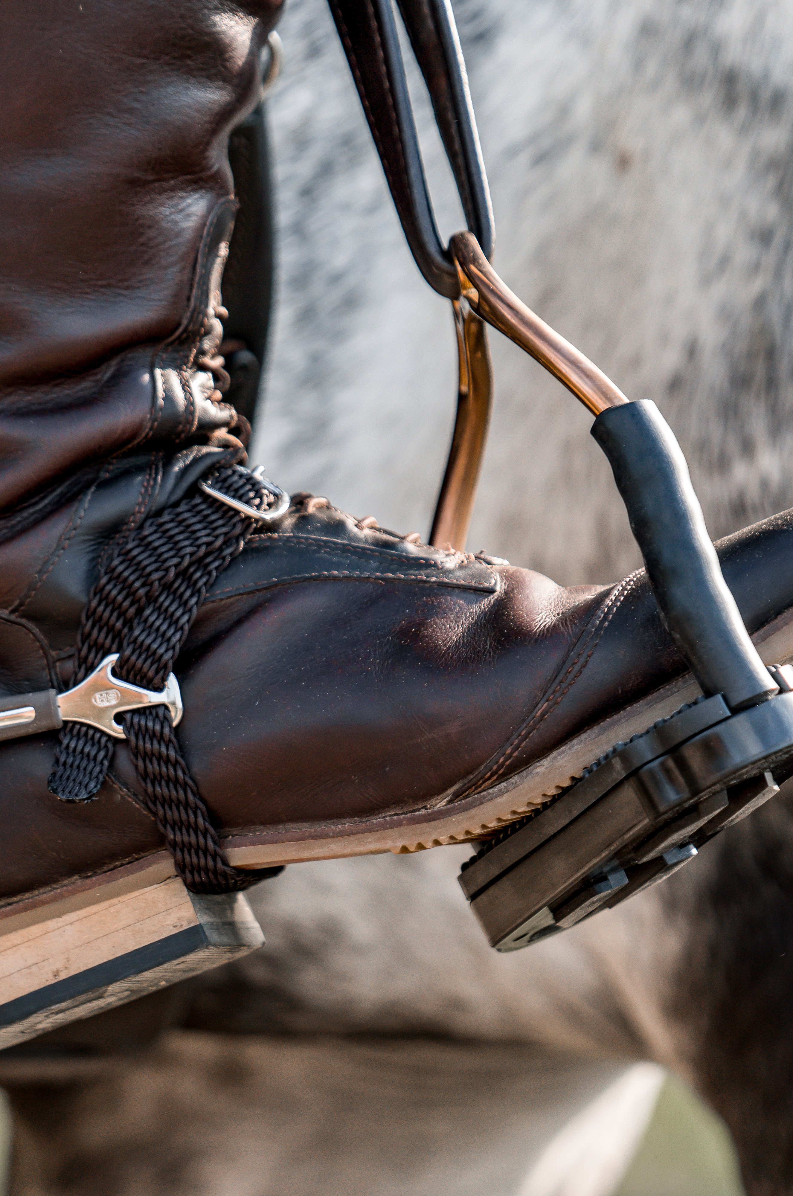 Ein detailgetreues Bild eines braunen Reitstiefels, der an einem Sattel und einer Steigbügel hängt. Der Stiefel ist aus Leder, mit einer Schnürung an der Vorderseite und einer Lederschnur um den Schaft, die sowohl Stil als auch Funktionalität bietet. Im Hintergrund sind unscharfe Konturen eines Pferdes zu sehen, was auf einen Reiteraktivität hindeutet. Die Sohle des Stiefels ist robust und hat eine klare Struktur, ideal für den Einsatz im Reitsport.